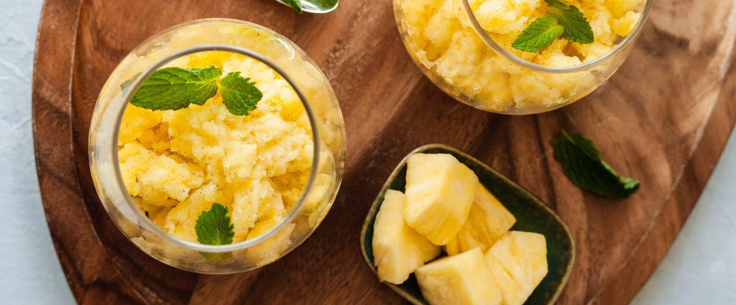 Pineapple-Vanilla Granita in two squat wine glasses on a wooden chopping board with a small bowl of pineapple chunks to the side