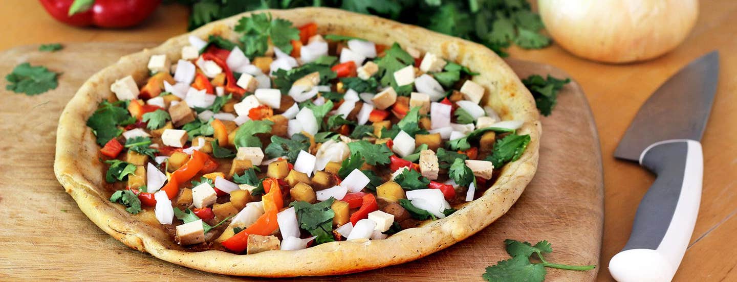 Vegan pizza topped with colorful chopped veggies atop a wooden chopping board with an onion and sprigs of parsley in the background