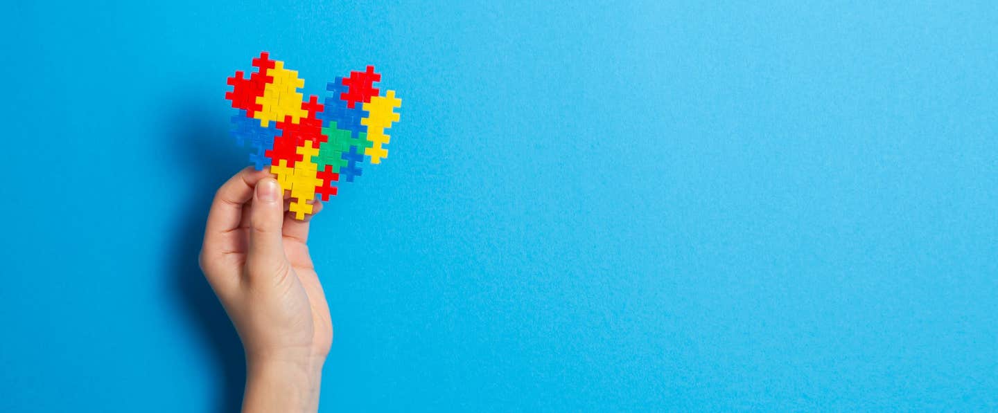 A hand holds up a heart made of colorful jigsaw pieces, against and blue background