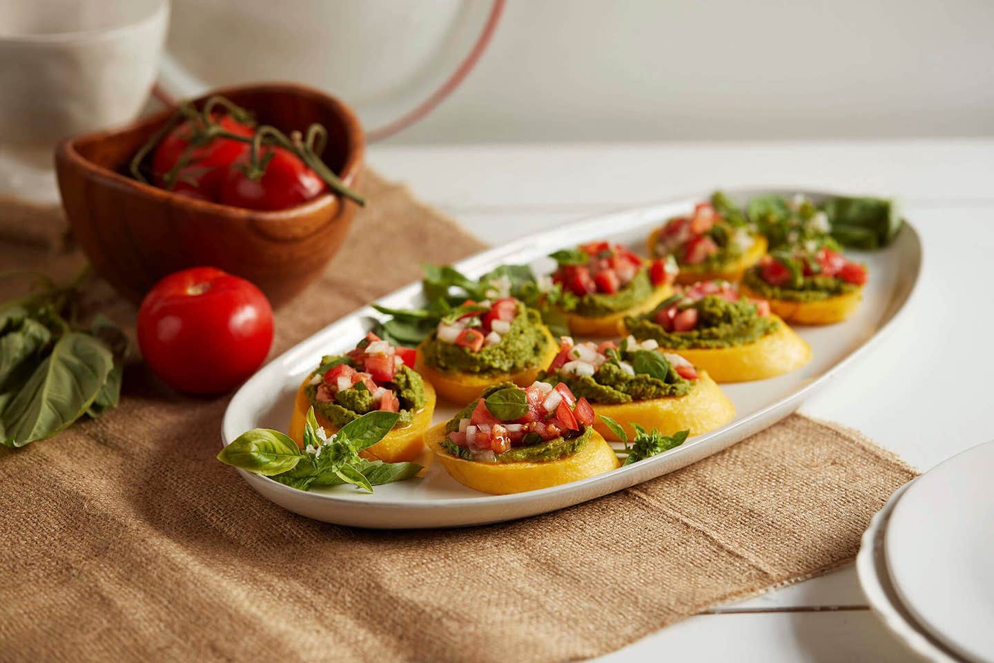 Polenta Crostini with Chickpea Pesto on a white oval serving platter on a hessian bag next to fresh basil and fresh tomatoes