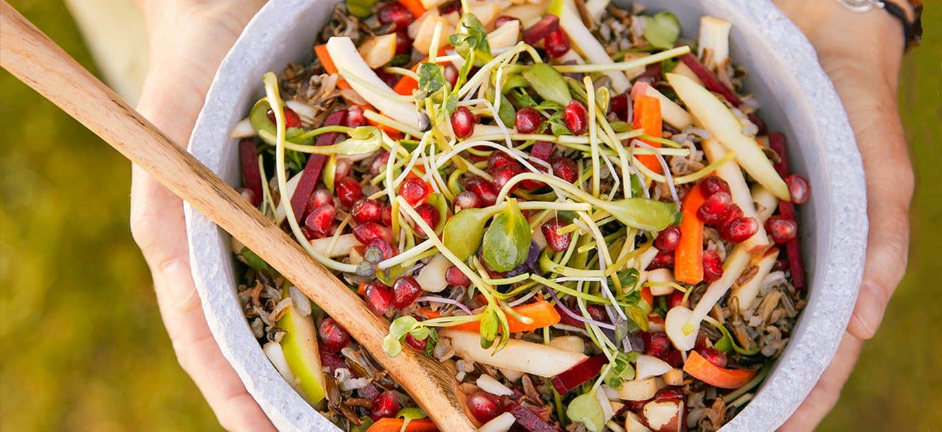 Hands holding a white marble bowl of Pomegranate Wild Rice Salad, with a wooden serving spoon in the bowl