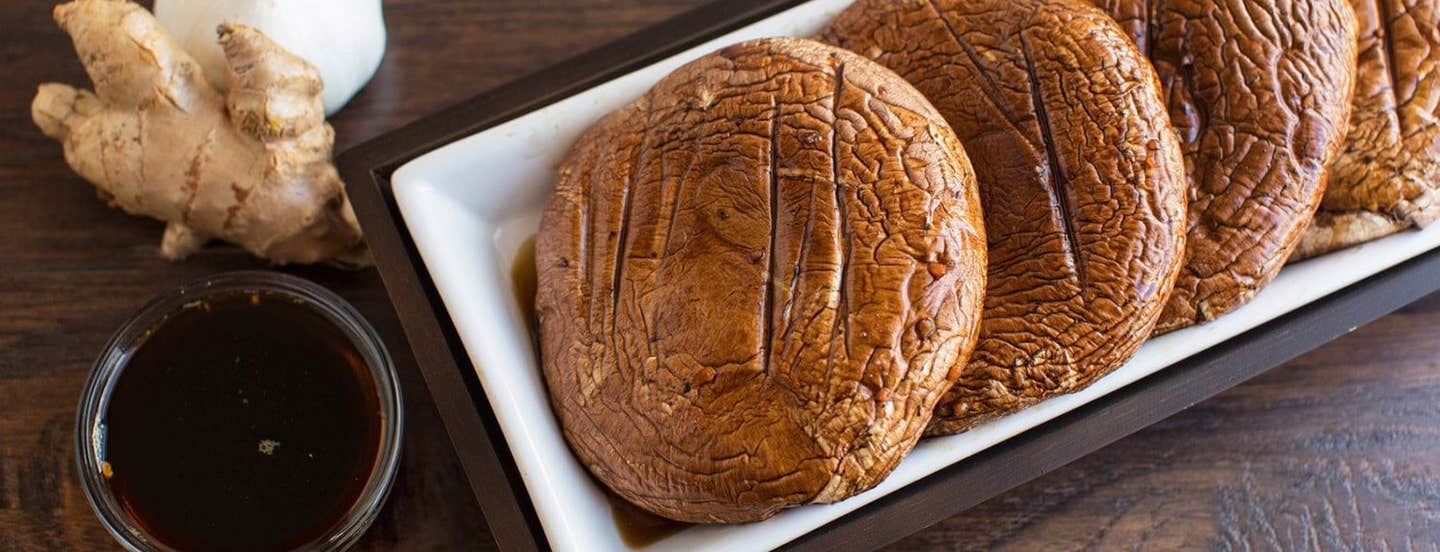 Four Grilled Portobello Mushrooms in a white rectangular dish next to a small bowl of marinade, a piece of fresh ginger, and a bulb of garlic