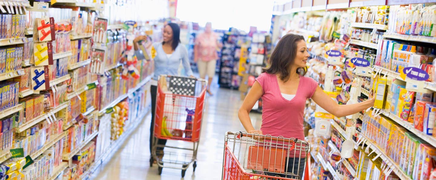 Looking down the cereal aisle of a supermarket. Two woman with shopping carts, reaching to grab a box of cereal