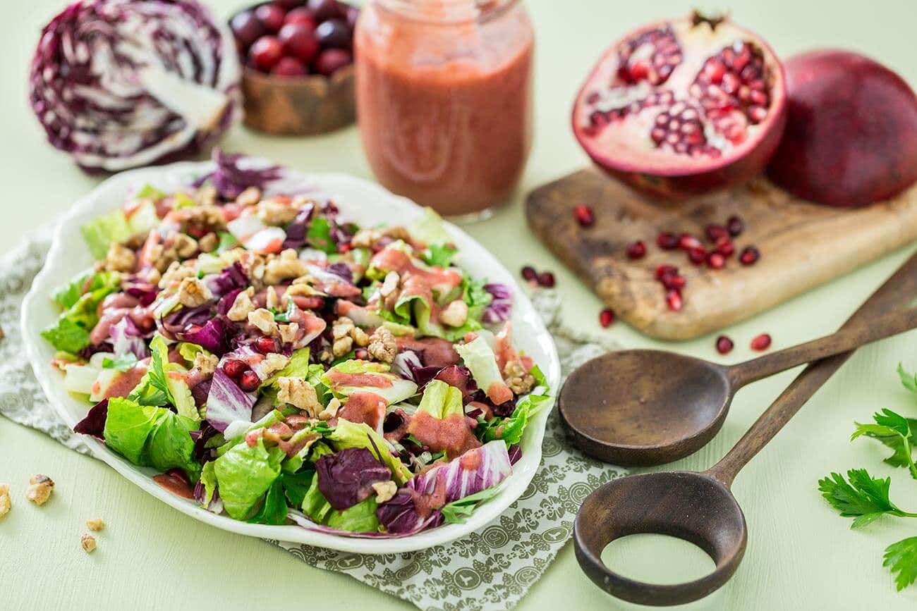 A large summery spread on a pale green table cloth, which includes a platter of Radicchio Salad with Cranberry Orange Dressing, wooden servings spoons, fresh pomegranates, and half a radicchio lettuce