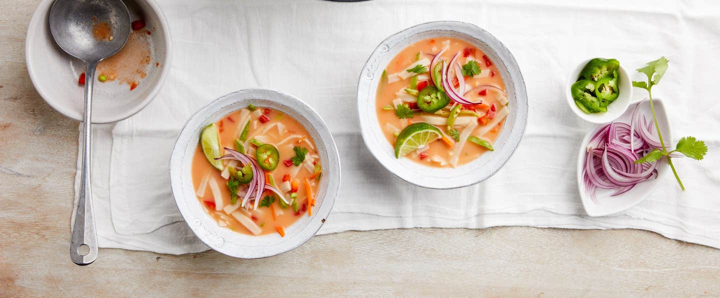 Two colorful bowls of Red Curry Noodle Soup on a white cloth with a small bowl of jalapeno and a bowl of sliced red onion.