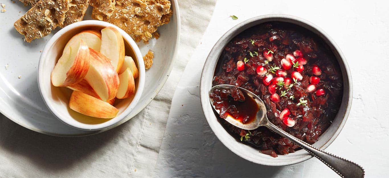 Red Onion Confit in a small gray bowl with a metal spoon next to a plate of crackers and apple slices