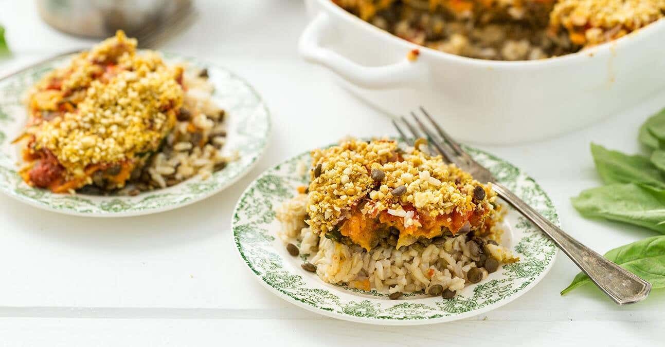 Two plates of Vegan Rice Casserole with Lentils, with a fork on the side. In the background is a large white casserole dish filled with the rice and lentil mix.