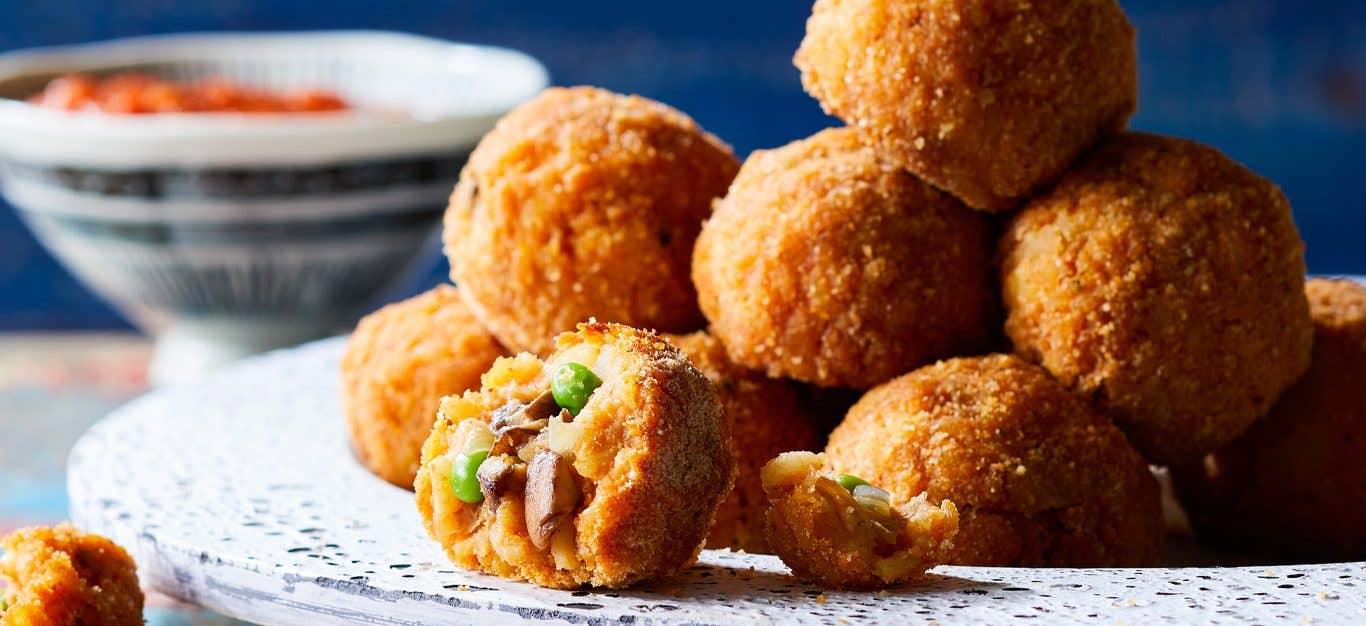 Sicilian Rice Balls with Mushrooms and Peas piled on a white platter with bowl of marinara sauce in background