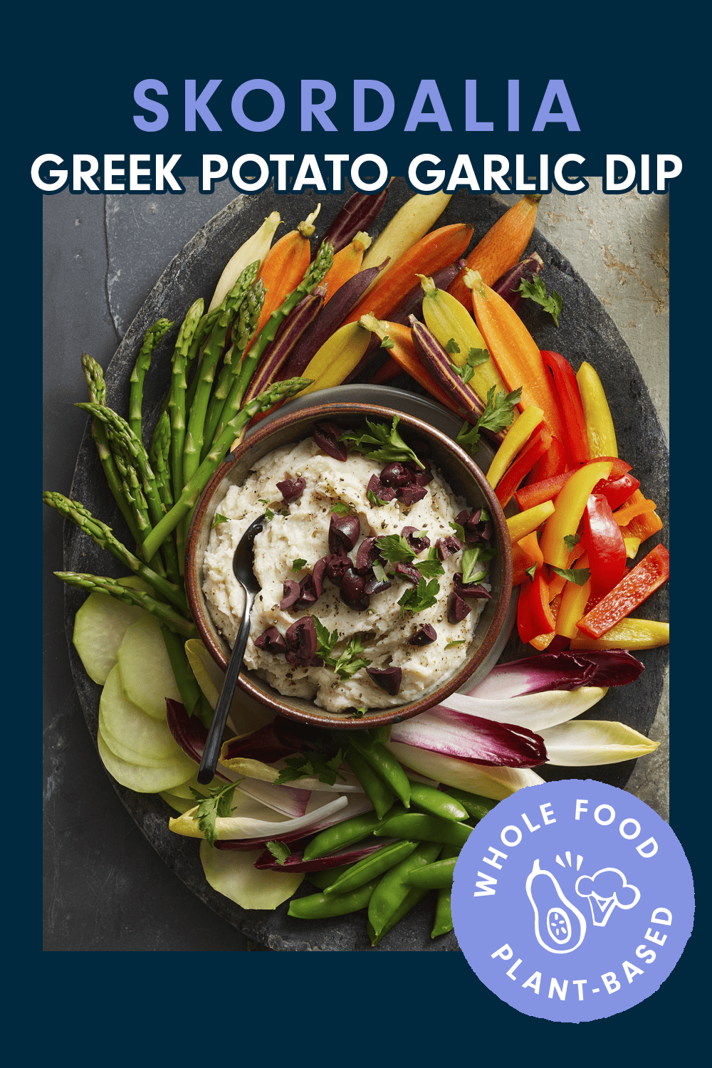 A bowl of dip on a platter surrounded with asparagus, baby carrots, radicchio lettuce, bell pepper, and sugar snap pease. Text reads, "Skordalia (Greek Potato Garlic Dip)"