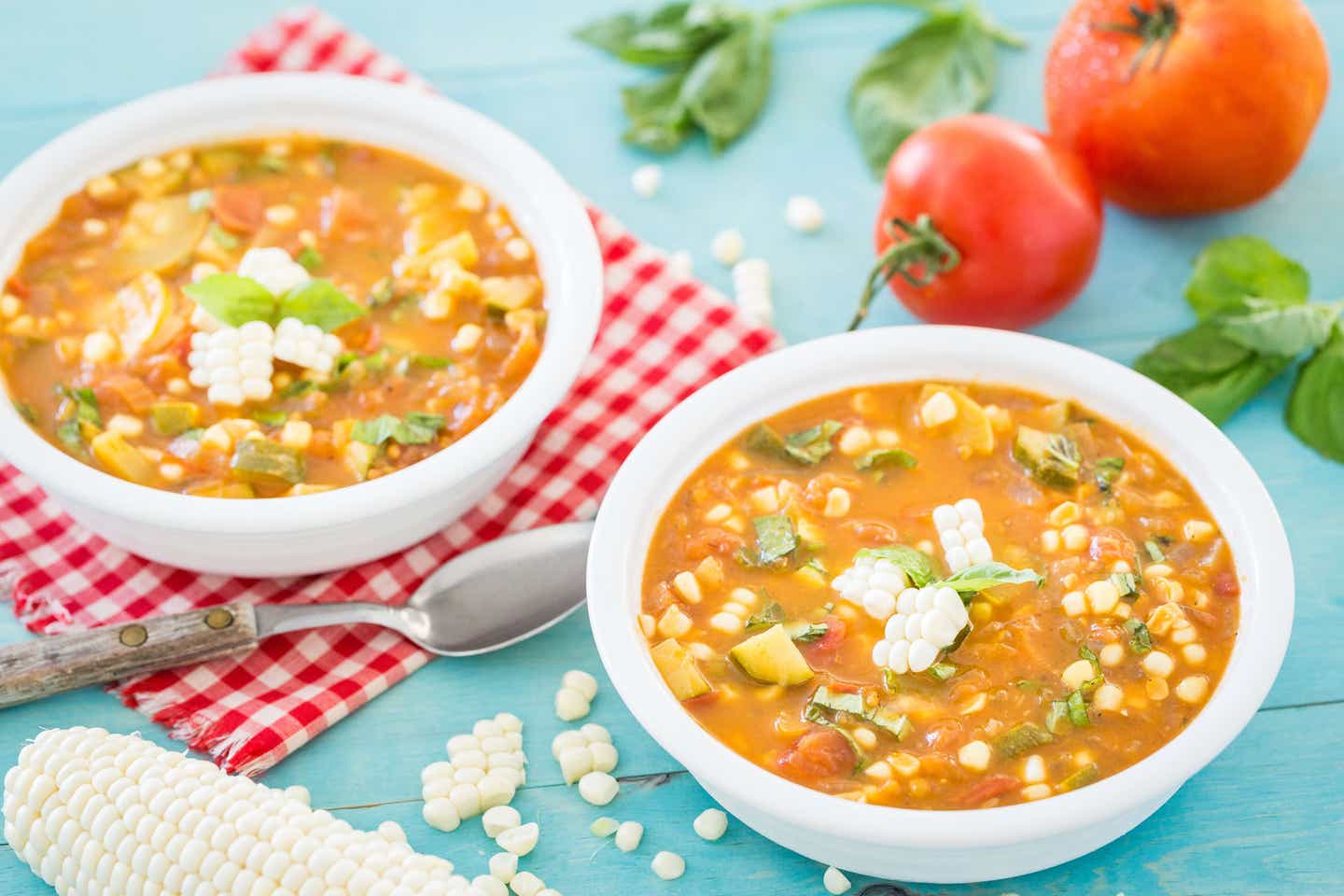 Two bowls of Summer Vegetable Soup garnished with fresh basil and planks of corn, on a red and white checked napkin and blue table. Fresh tomatoes, corn, and basil are in the background.