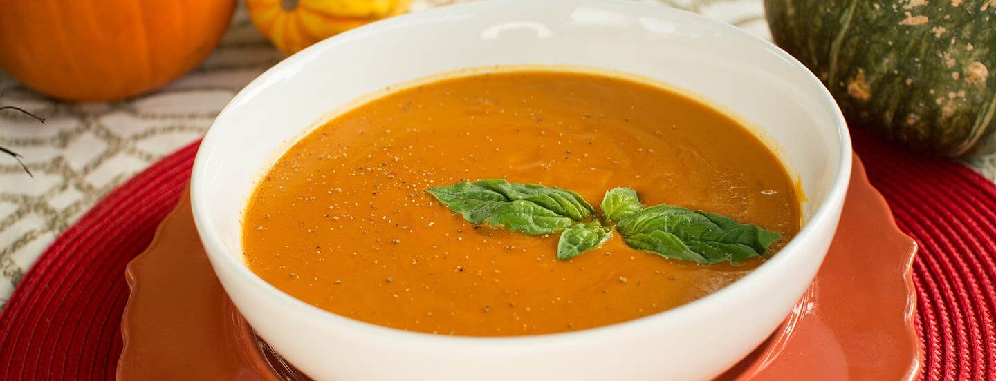 A white bowl of Sweet-Potato-Bisque on a red plate, with a sprig of fresh basil on top