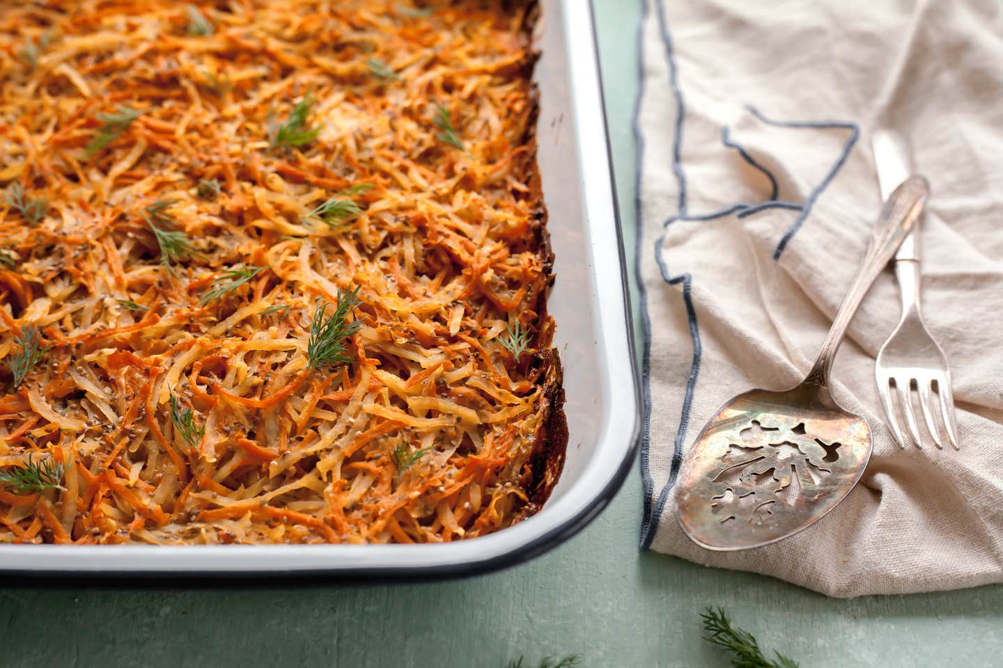 A casserole dish of Sweet Potato Kugel next to a white kitchen towel and silver serving spoons