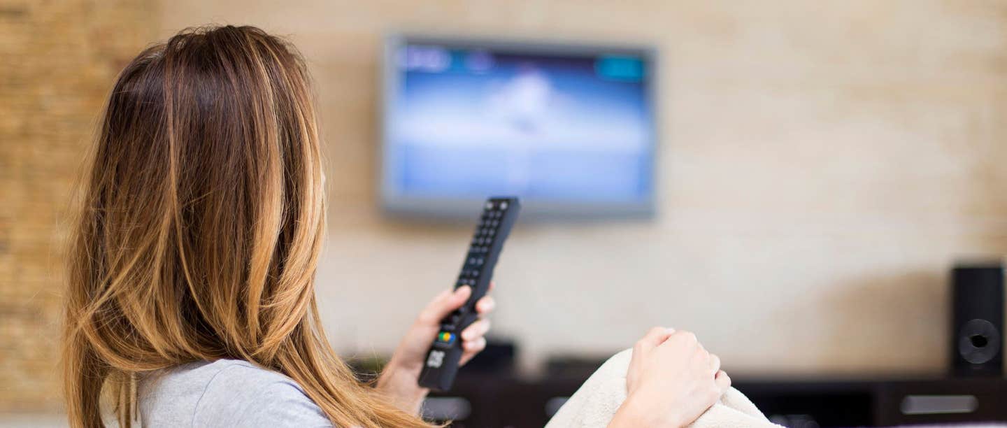 The back view of a woman holding a remove looking at TV screen in the distance