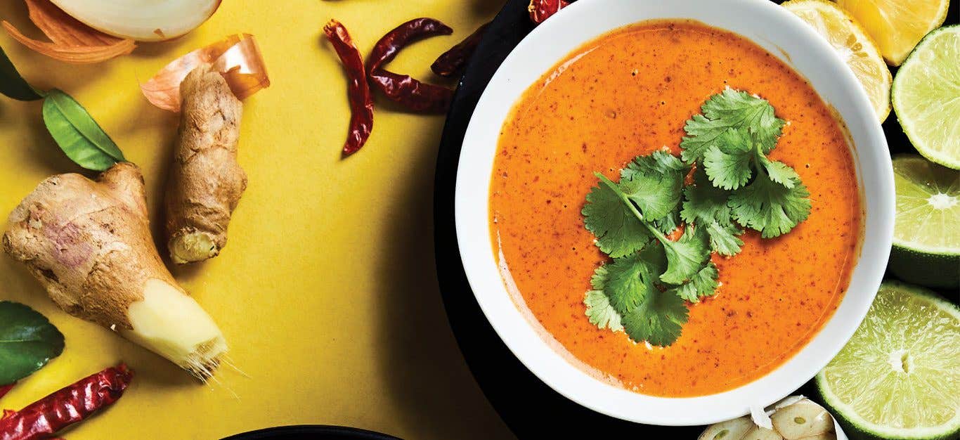 Thai Red Curry Sauce in a white bowl against a yellow background surrounded by lime slices, ginger root, and dried chili peppers