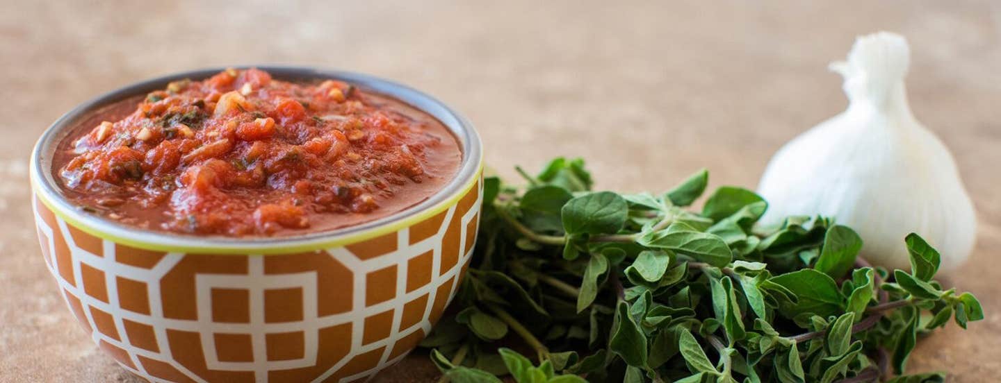 Tomato Sauce in a decorative brown ceramic bowl next to a bunch of fresh herbs and a head of garlic.