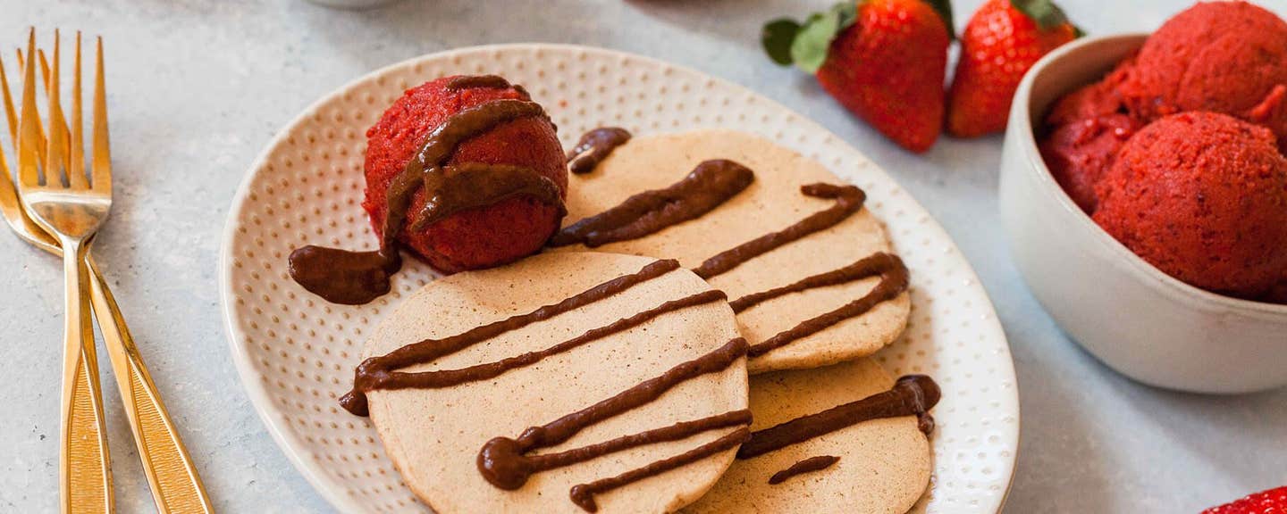 Vanilla Pancakes with a scoop of Strawberry-Cherry Ice Cream drizzled with Warm Chocolate Sauce. In the background is a bowl of chocolate sauce, a bowl of ice cream, and fresh strawberries