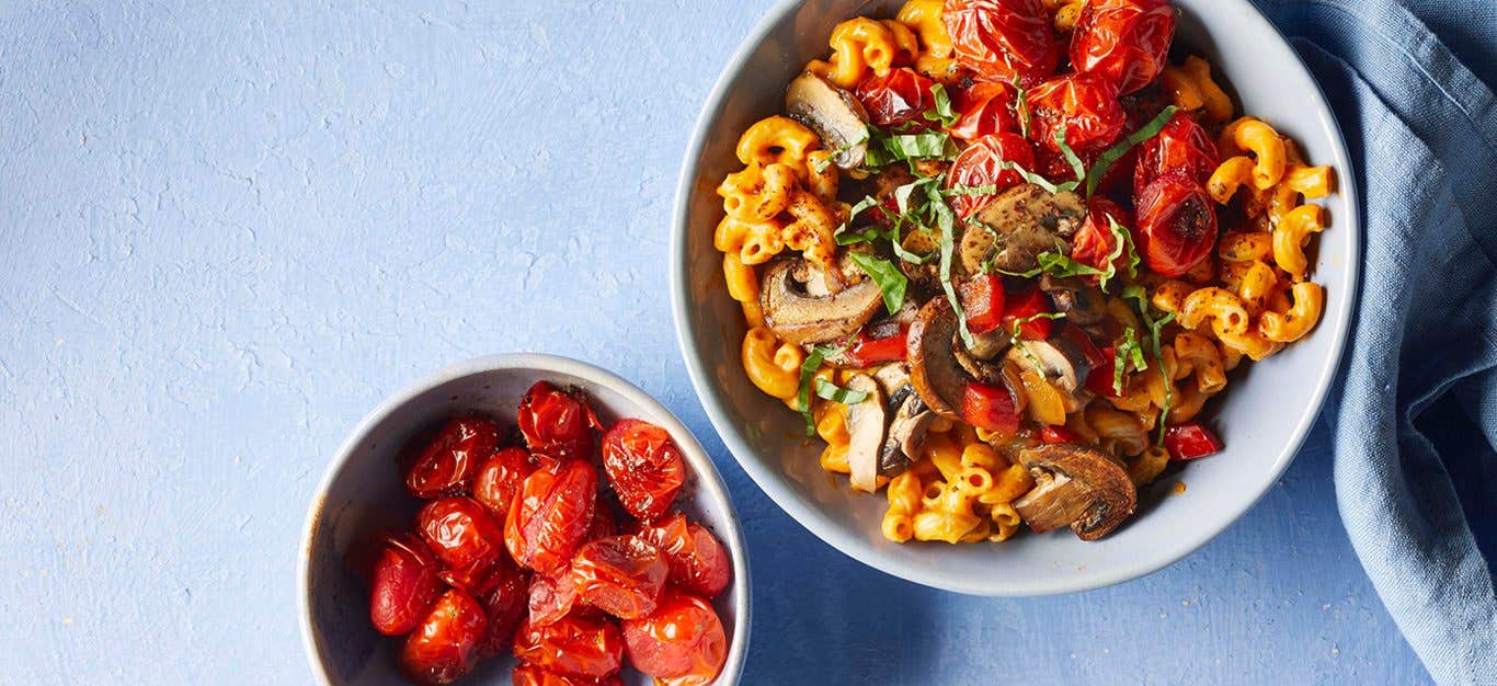 Vegan Mac and Cheese with Queso against a blue background next to a small bowl of roasted cherry tomatoes