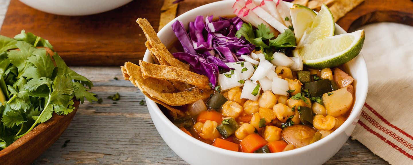 A white serving bowl Vegan Pozole garnished with lime wedges, tortilla strips, finely chopped red cabbage and onion