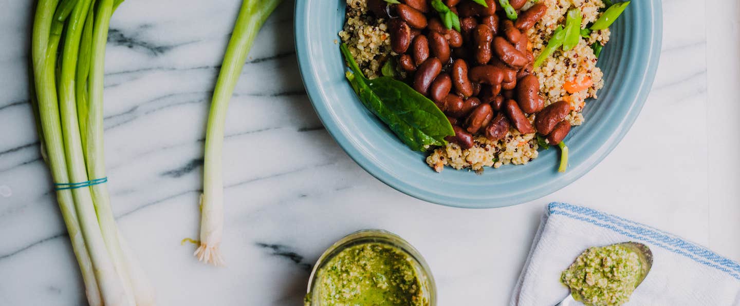 A blue bowl of quinoa, kidney beans, and greens on a marble couner next to green onions and a small bowl of green sauce