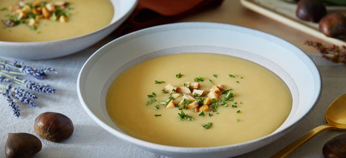 A wide-rimmed pale grey bowl of Winter Potato Leek Soup. Next to the bowl are chestnuts and dried rosemary