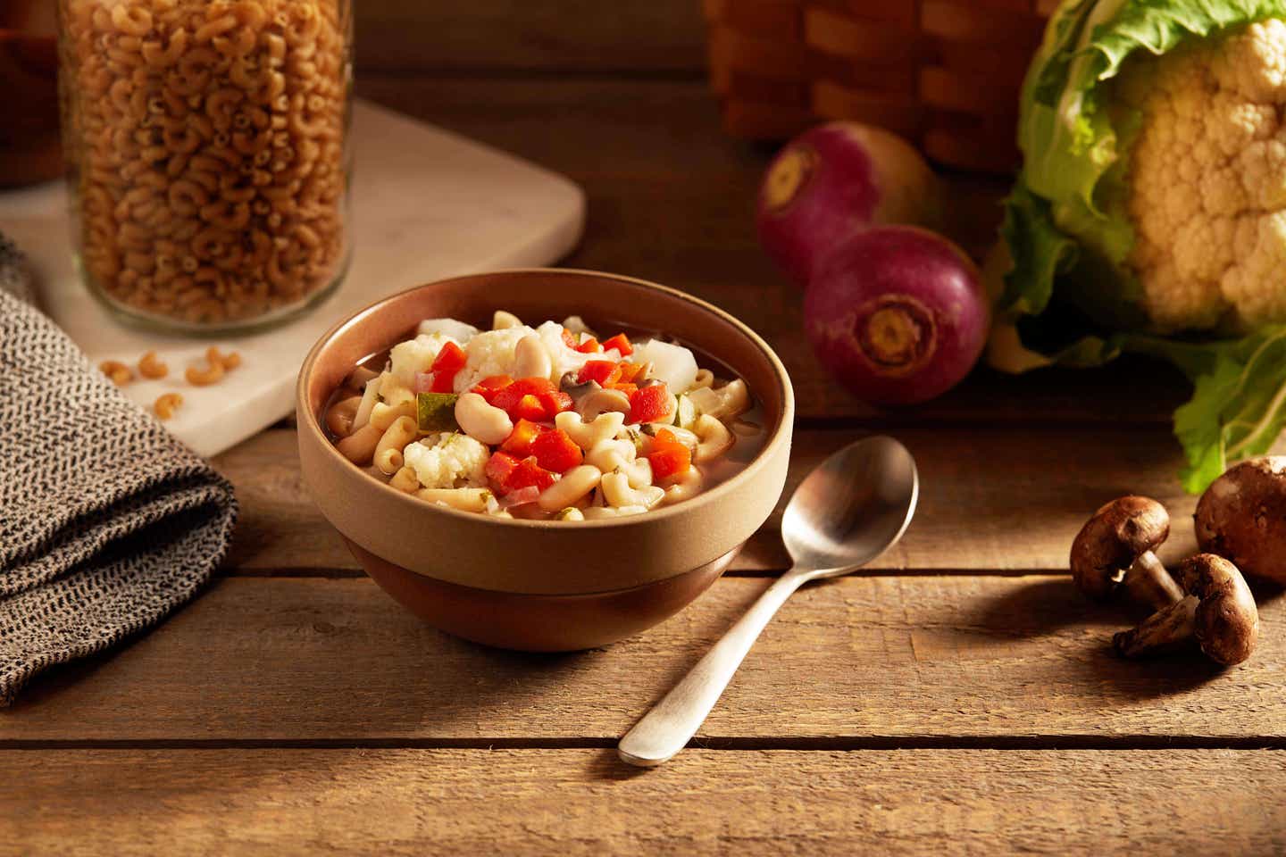 Winter White Bean Minestrone Soup in a brown ceramic bowl on a rustic wooden table top with raw vegetables and a jar of dried macaroni