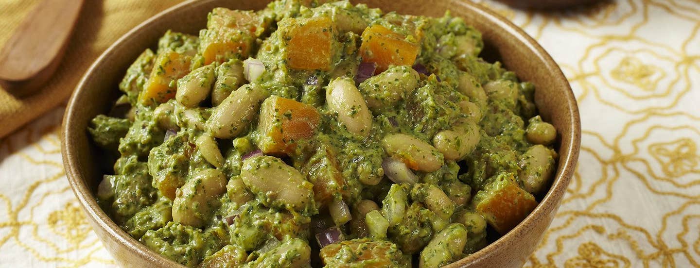 A brown ceramic bowl of Winter White Bean Salad with Spicy Cilantro Pesto. In the background is a wooden serving spoon and a smaller wooden bowl of pesto