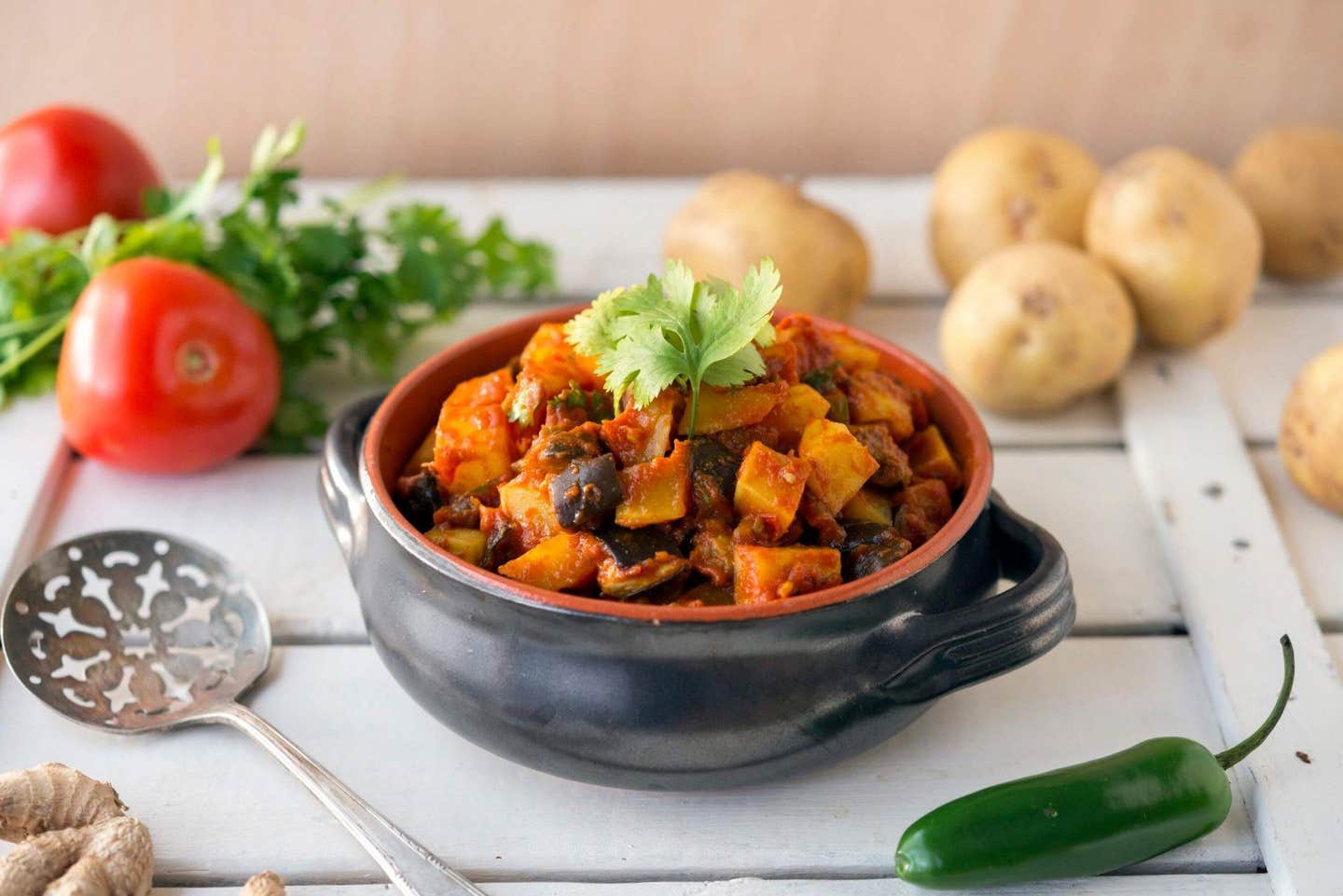 A soup with handless, filled with a hearty stew: Aloo Baingan (Potato and Eggplant). in the background are raw potatoes, tomatoes, parsley, and chili