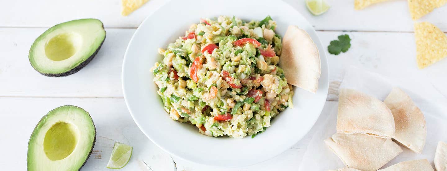 Smashed Chickpea Avocado Dip in a white bowl on a white background with pita bread squares and a whole avocado cut in half off to the side