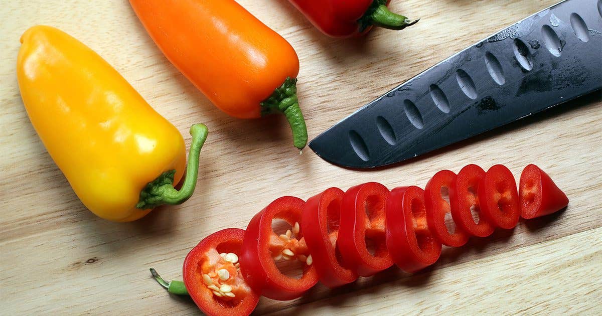 A close-up of a wooden chopping board with colorful peppers, a knife blade, and finely sliced red chile