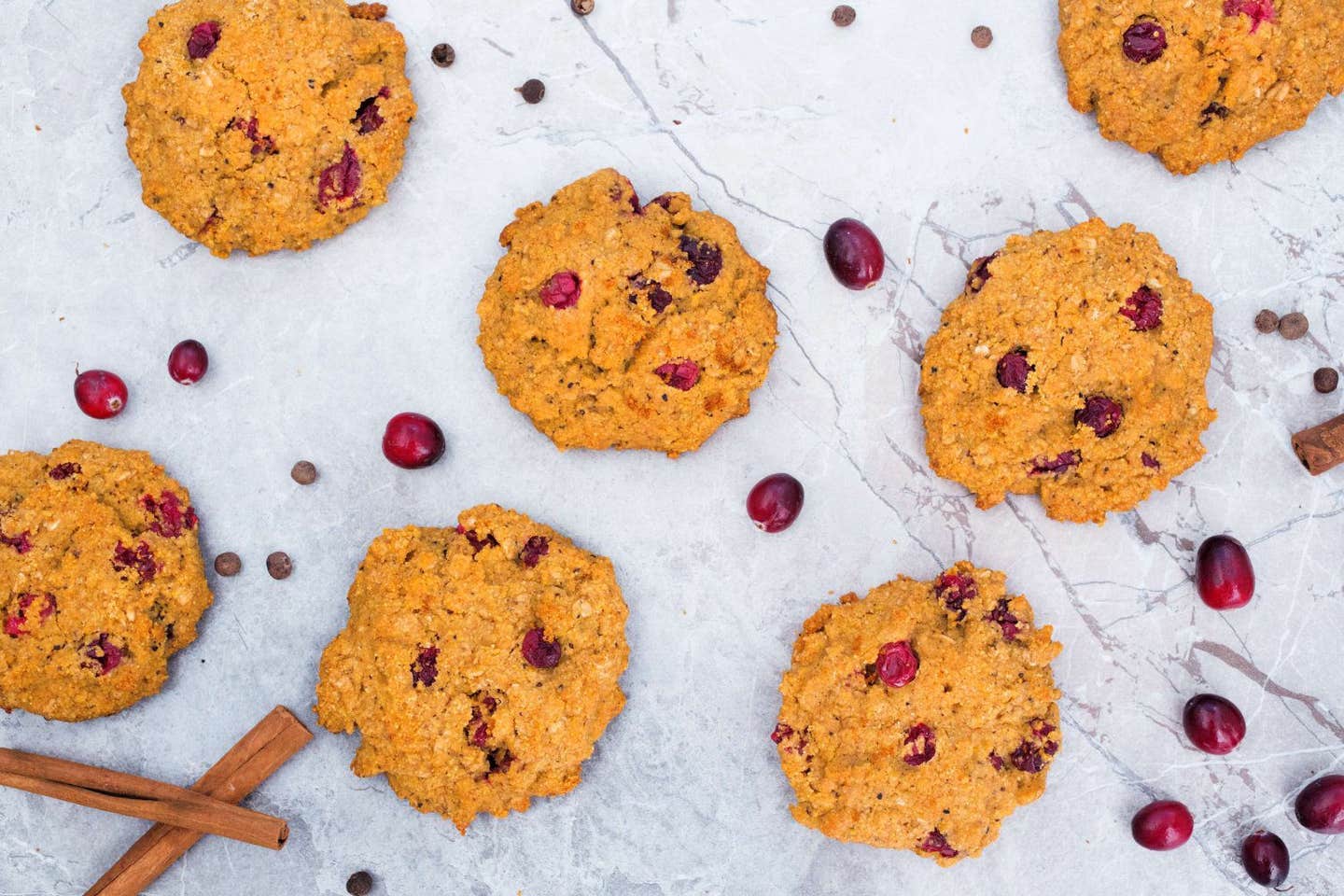 Pumpkin Cranberry Cookies spread out on a marble background with cinnamon sticks and fresh cranberries scattered between the cookies