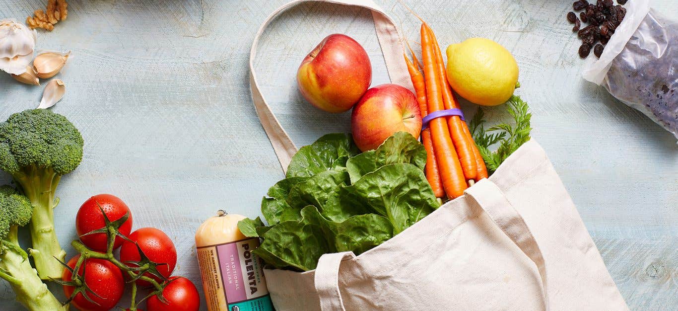 A canvas tote bag full of fresh produce lies on a white counter with more veggies surrounding it