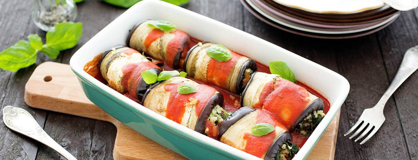 A casserole dish atop a wooden chopping board, filling with six Eggplant Rollatini drizzled in a tomato sauce and garnished with a leaf of fresh basil