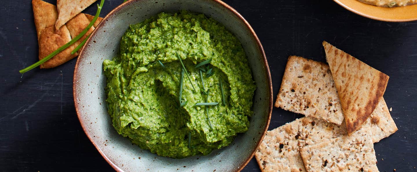 Emerald Hummus Dip with Fresh Herbs garnished with chives, with whole grain crackers around the bowl