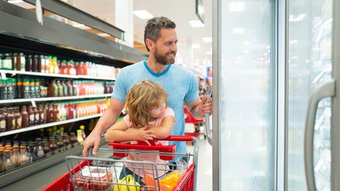 Smiling man opens the door of a refrigerated section at the grocery store, while his young daughter sitting in the cart watches and smiles