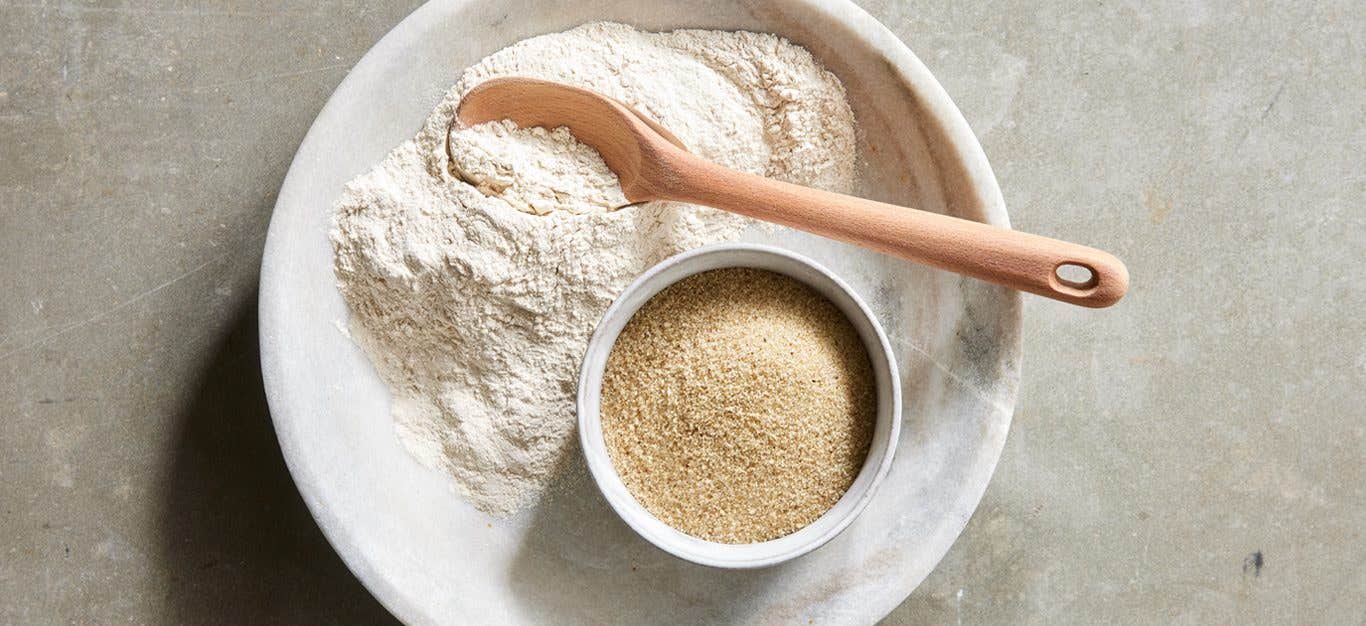 A flat-lay shot of a plate with some ground fonio flour on it, a wooden spoon, and a small bowl with fonio grains in it