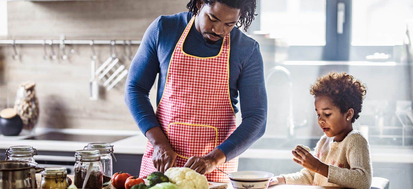 Black father and daughter in kitchen chopping vegetables, with daughter examining a piece of vegetable - teaching children about cultures through food