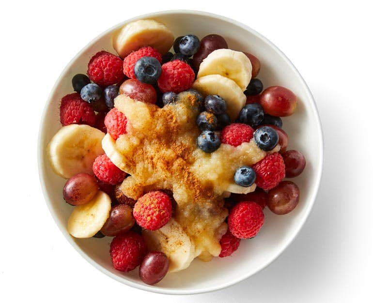 Chopped bananas, berries, an grapes ion a white bowl with applesauce and cinnamon on top