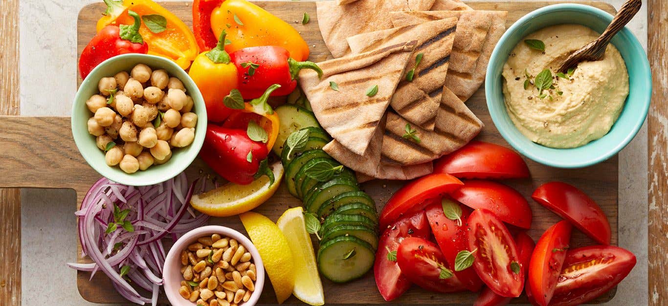 greek salad board with chopped veggies, pita bread, and bowls of hummus on a wooden serving board