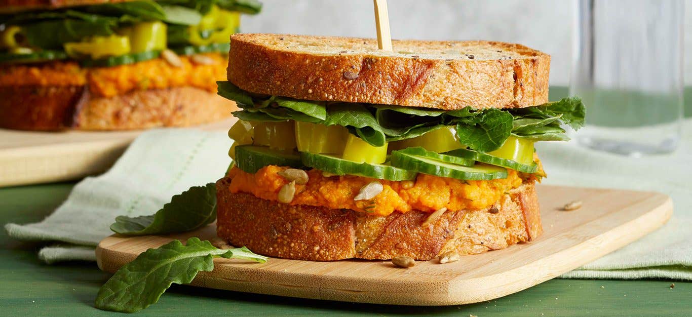 Close-up shot of a vegan sandwich with homemade hummus, pepperoncinis, and cucumbers, stacked between two pieces of whole grain bread