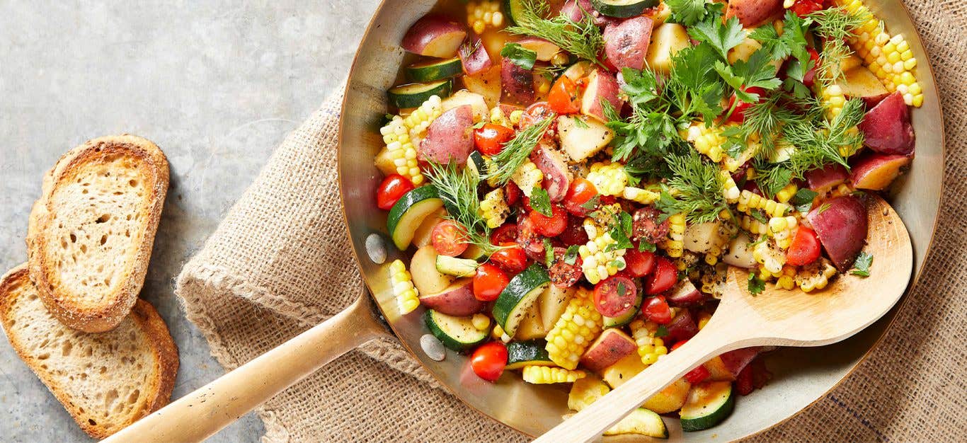 herbed breakfast hash in a metalskillet with a wooden spoon and slices of toasted bread on the side