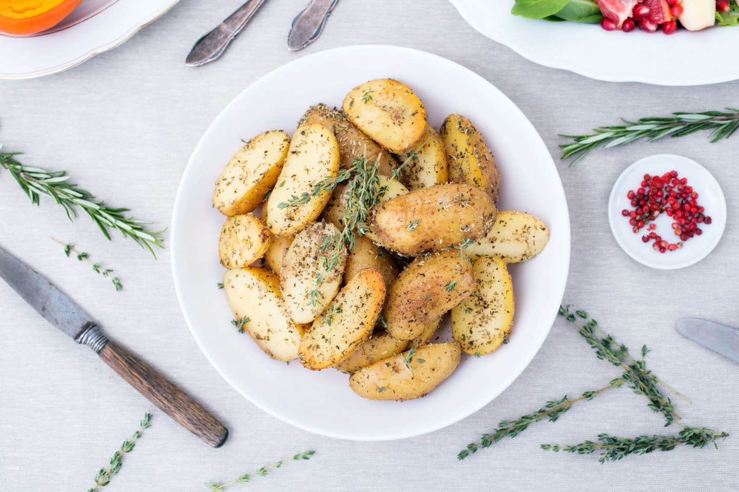 Small, herb-coated, cooked potatoes cut in half presented on a white plate with a sprig of rosemary on top. The plate sits on a marble table top with more rosemary and a small bowl of pomegranate arils beside it.