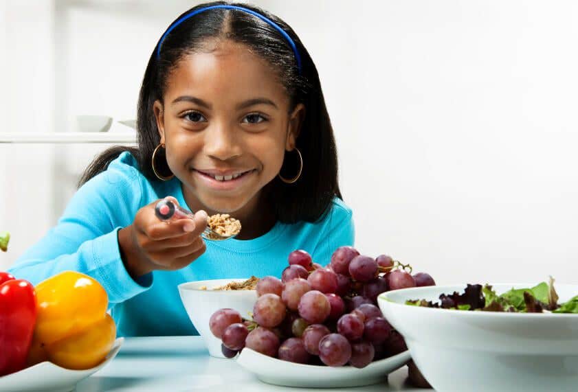 young girl sitting at a table eating healthy food, in front of a bowl of grapes and a bowl of greens