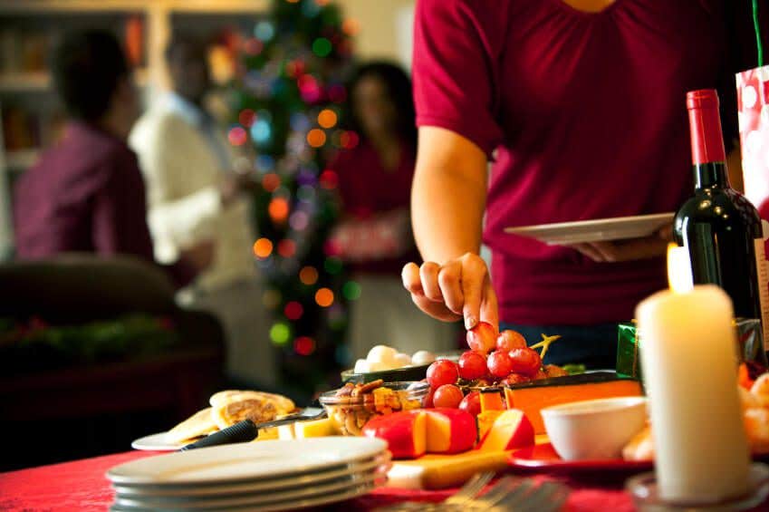 A hand reaches out to grab a grape from a holiday table which also has a block of cheese, a candle, and a bottle of wine