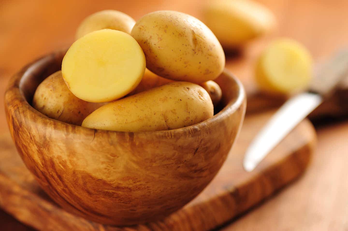 A wooden bowl of raw potatoes with a knife in the background