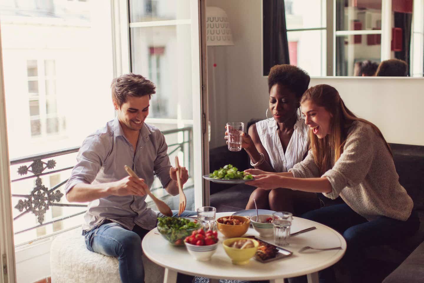 Three young adults laughing and sitting around a small table, sharing healthy plant-based food