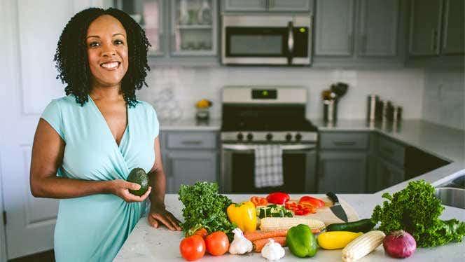 Doctor Judy Brangman holding an avocado in a kitchen with lots of fresh produce on the counter