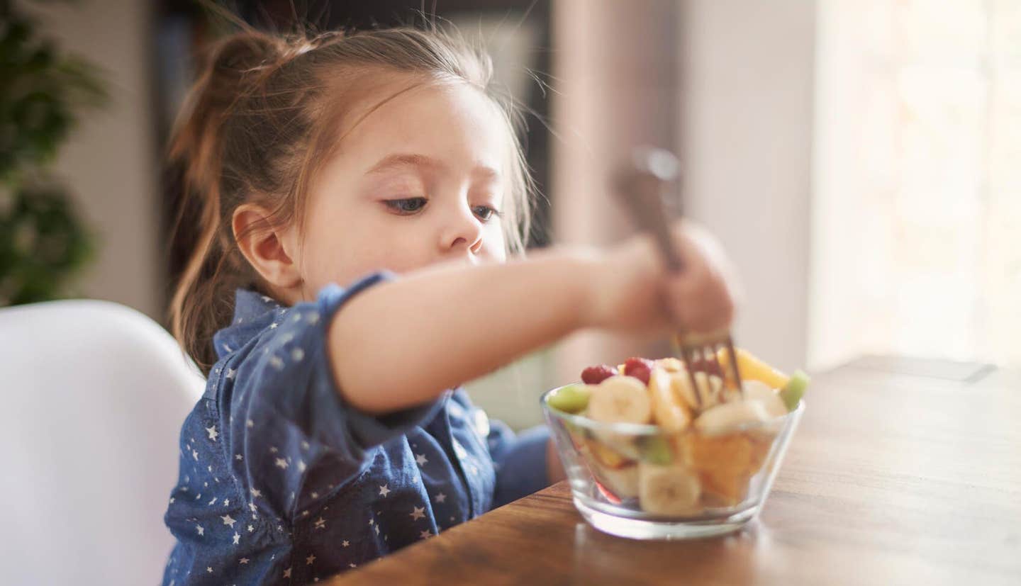 A two-year old girl at the table eating fruit salad with a fork