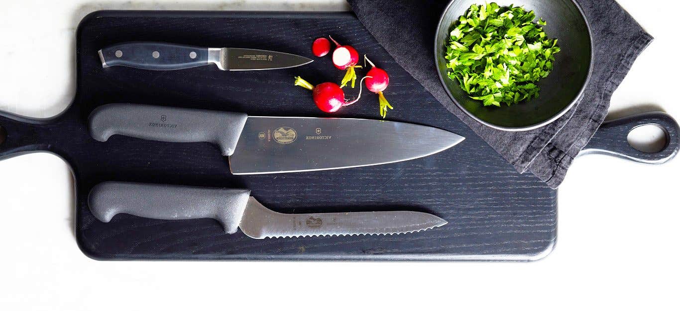 A dark cutting board with three knives laid out parallel across the board, along with a few radishes and a small bowl with finely chopped herbs