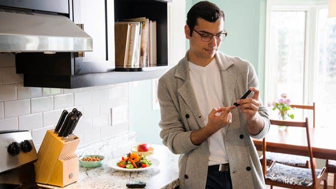 man with type 2 diabetes tests blood sugar while standing beside his kitchen counter, where a plate of vegetables sits