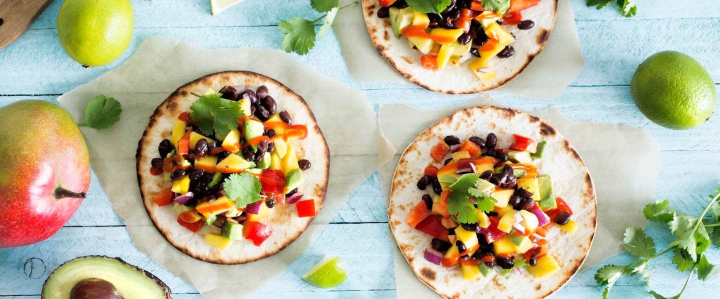 Three Mango and Black Bean Tacos, each on a small piece of parchment paper, against a pale blue wooden background, with fresh mango, avocado, and lime dotted around them